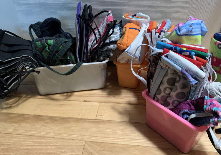 Image shows a white rectangular plastic container holding large masks. The left is more black and the right more colorful. The bins and masks shown in the previous photo are placed on the right side of this image beside the white container.