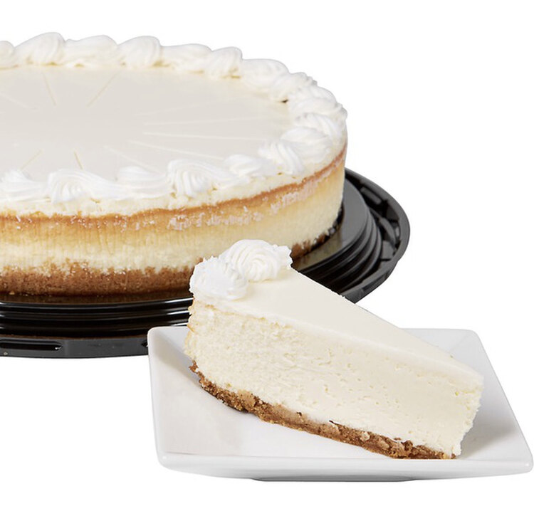 Image shows a whole cheesecake on the black plastic base without the clean lid overtop. The left side is cut off and in front of it, on a white square plate, sits an unadorned slice of cheesecake.