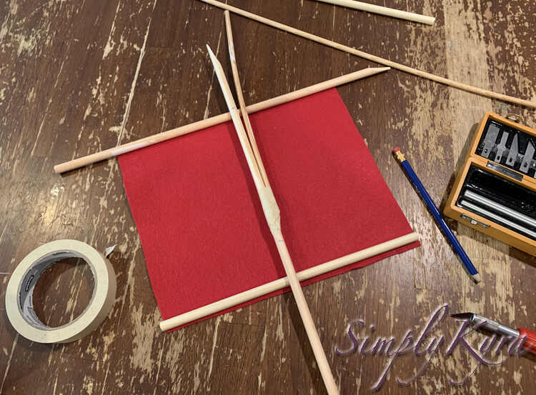 Image shows the sail laying flat on the table with the mast pieces laid overtop. The main vertical mast now has a branch curving away and taped at it's base. There's a mark on the vertical mast where the top horizontal beam is and another mark about an inch above the bottom beam.