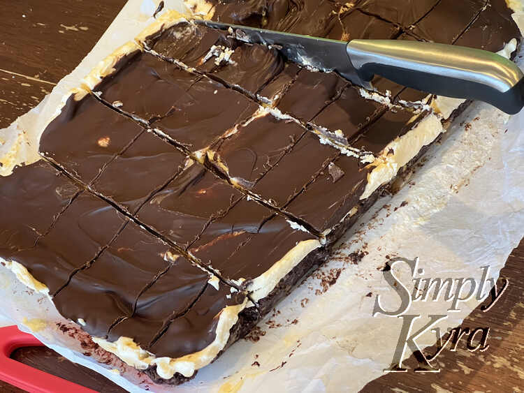 Image shows the scored Nanaimo bars laid out on a cutting board with a knife cutting the pieces apart.