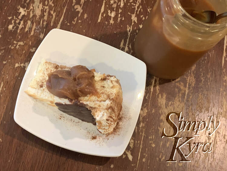 Image is taken looking down at a slice on its side with a heap of caramel on top and a sprinkling of cinnamon showing. To the side is a large jar with cold caramel and a spoon inside to dish.