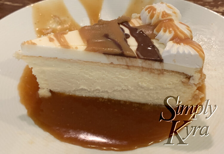 Image shows the hardened chocolate and caramel topped cheesecake slice sitting in a pool of hot caramel on a white saucer.