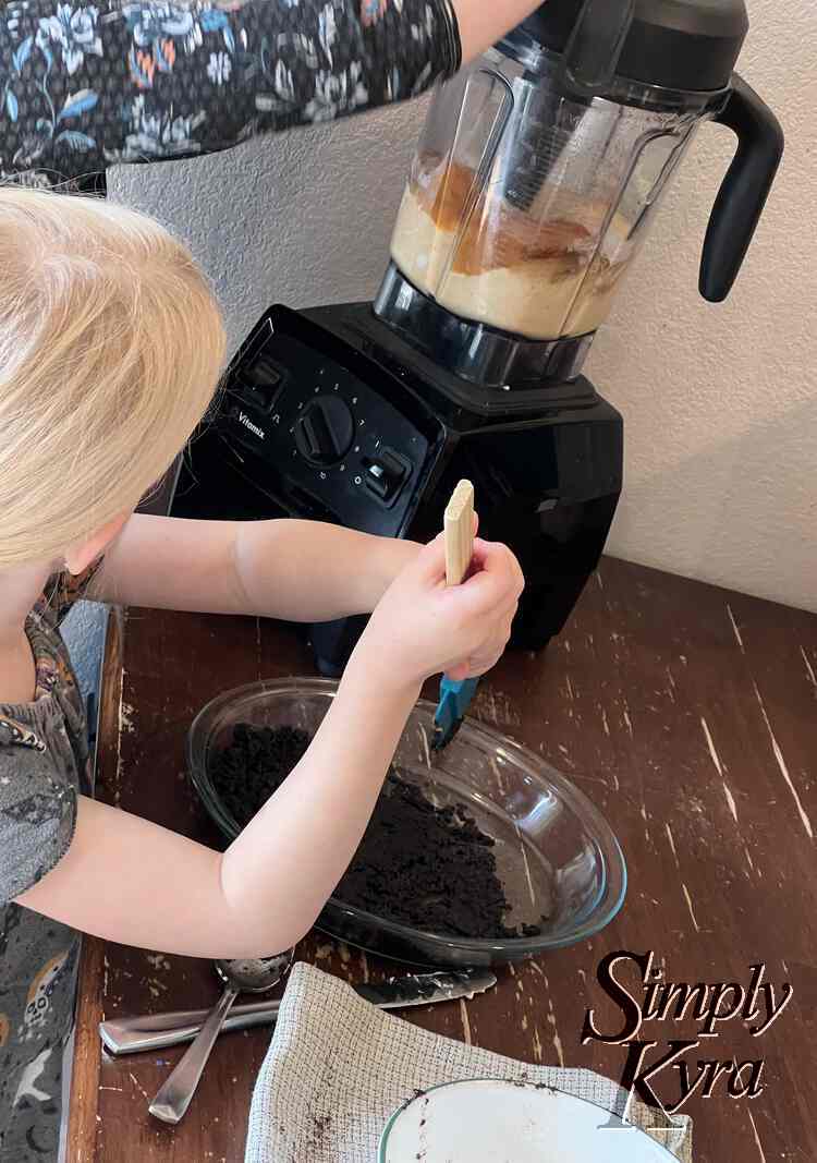 Image shows Zoey in the foreground using a blue spatular to press the chocolate crust into the glass pie plate. She's looking into the background at Ada and the blender as all the ingredients are mixed together.