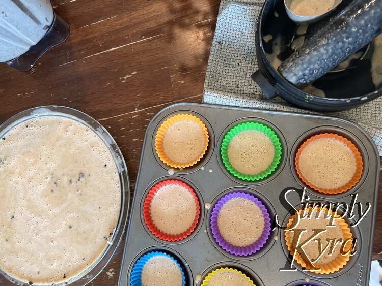 Image shows a glass pie plate filled with bubbly liquid to the left. To the right you can see six and a bit colorful cupcake liners half filled with more cheesecake mixture. To the top you can see the blender, on the left, and the blender accessories, measuring cup, and tea towel on the right.