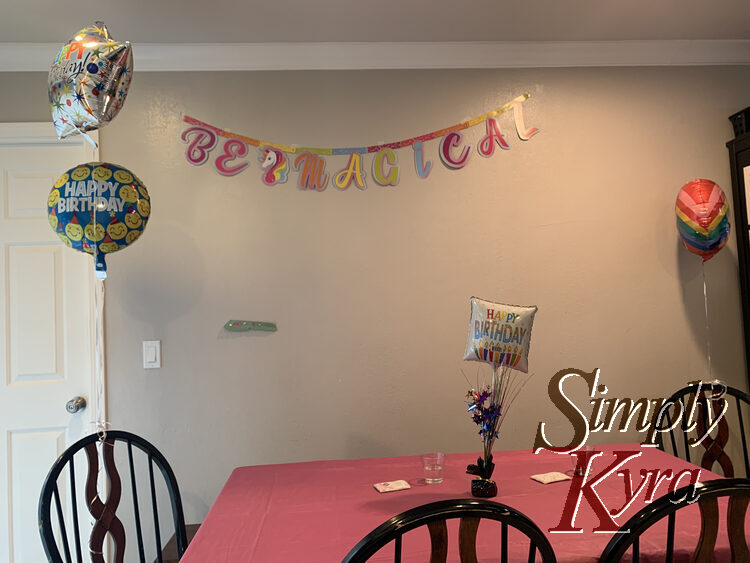 Image shows a pink tablecloth covered table with a Happy Birthday centerpiece, two happy birthday balloons on the left chair, a rainbow balloon on the right, and a "be magical" banner on the wall behind.