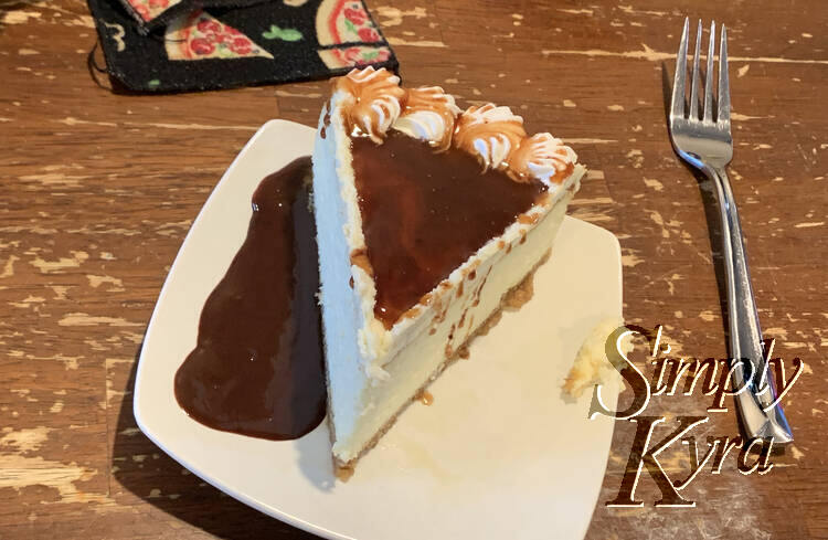 Image shows a slice of cheesecake from above with chocolate sauce covering the bit of caramel on and beside the slice. A fork and part of a pizza adorned napkin are laid out behind and beside the saucer.