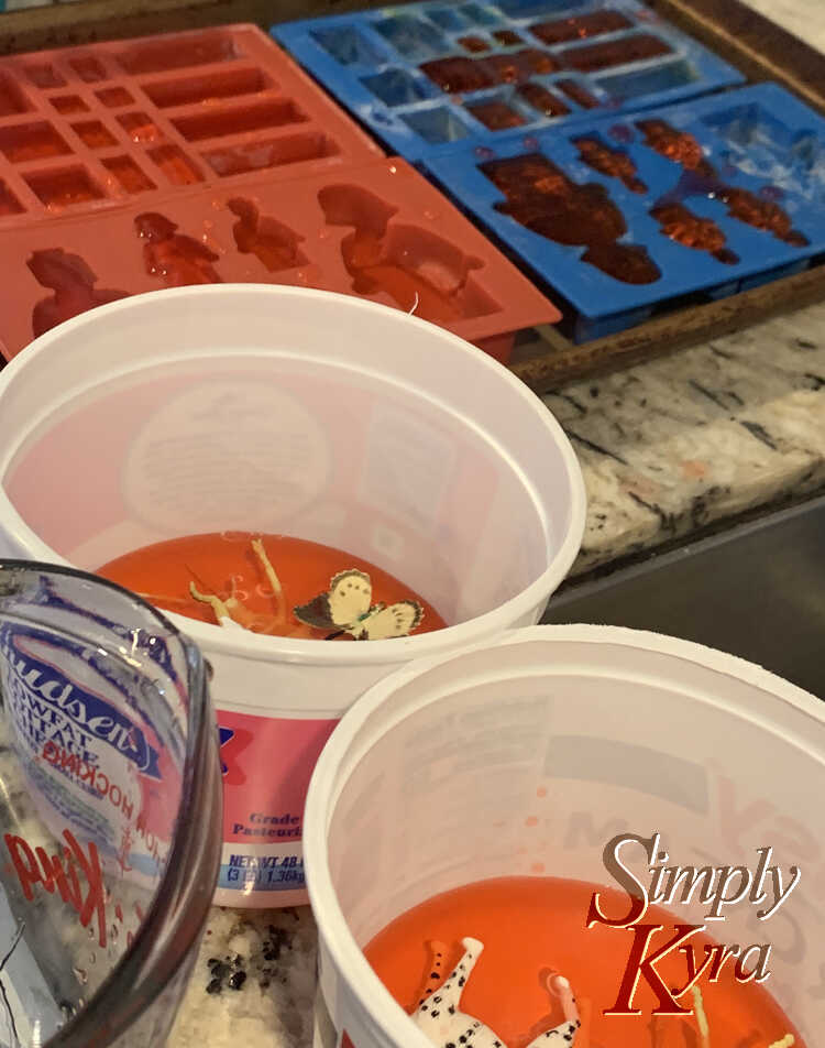 Image shows two plastic containers with plastic toys and orange water at the bottom. A glass measuring jug sits beside it. In the back are LEGO inspired silicon molds resting on a cookie sheet to be easier to move while filled with the orange water.