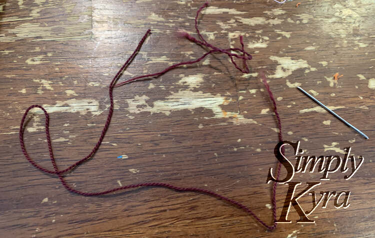 Image shows a reddish brown thread on a table with a sewing needle nearby. The end of the thread is frayed away with bits scattered around in front of it.