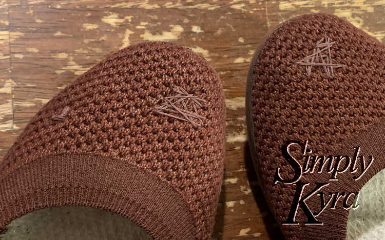 Image shows the front top of both shoes side by side over the brown table. They both have a decorative triangular or heart-ish criss-crossing design over their main hole. The left shoes also has a small left covering a little patch on the top side.