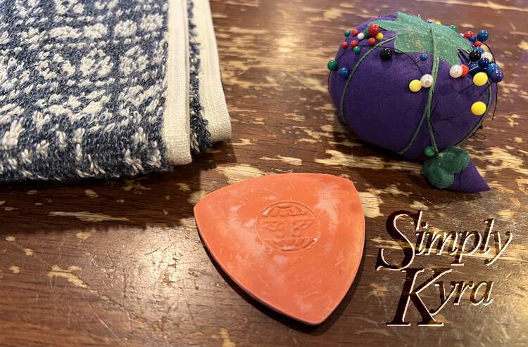 Image shows the edge of the folded towel beside a triangle of red tailor chalk and purple tomato with multi-colored sewing pins in it. 