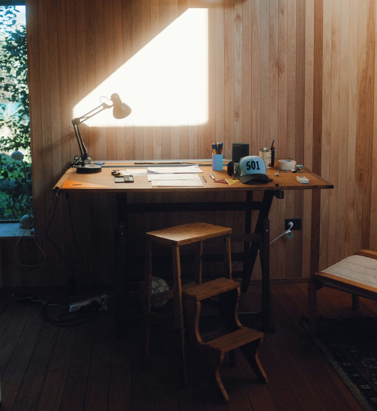A quiet creative studio with a wooden table, chair, table lamp, and a window view—capturing a lived-in, reflective workspace.