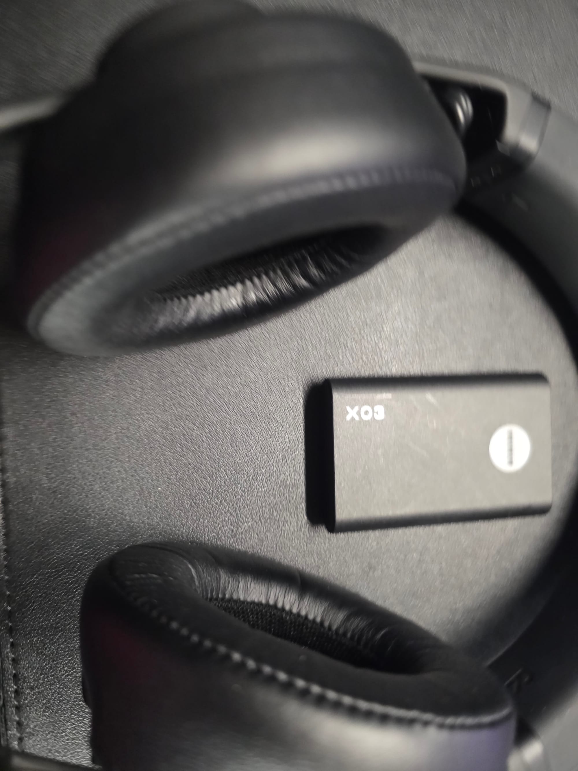 A close look at the AIAIAI TMA-2 studio headphones, which are black with black earcups.  The X03 transmitter is also black.  Both are lying on a black desk.