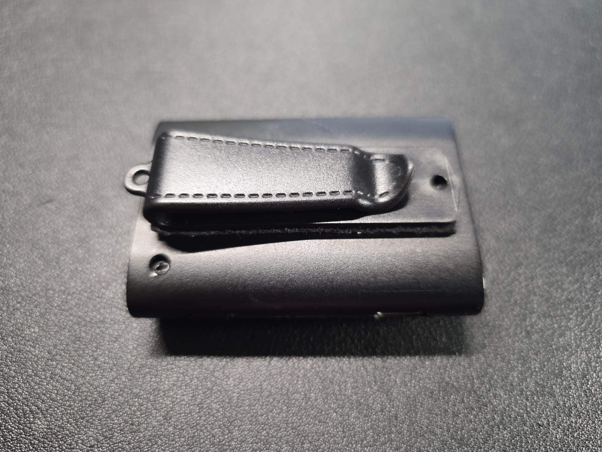 An Avantree receiver with the clip attached.  The clip has a hole for a lanyard at the top.  The receiver is lying face down on a black desk.