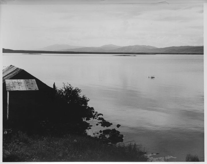 black and white photograph of a still lake, with a dark wood building on the shore