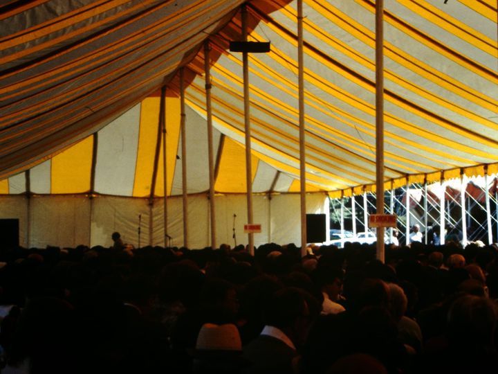 inside of a blue and yellow striped tent, a seated crowd is gathered