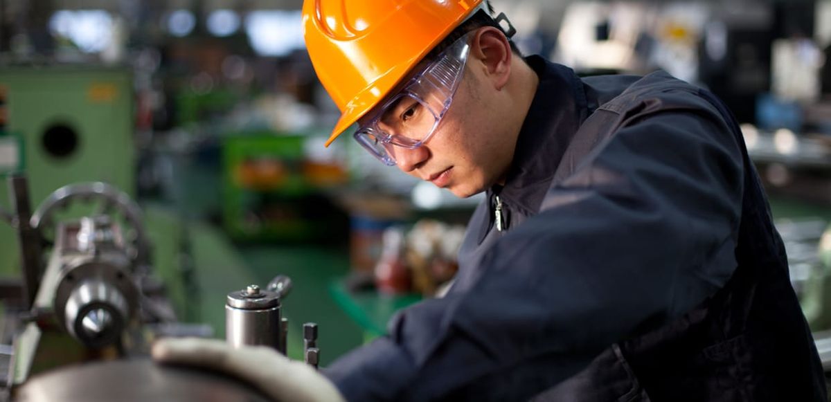 A factory worker wearing safety glasses, and orange hard hat, and a deep blue work uniform rests his hand on and inspects a piece of machinery