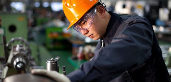 A factory worker wearing safety glasses, and orange hard hat, and a deep blue work uniform rests his hand on and inspects a piece of machinery