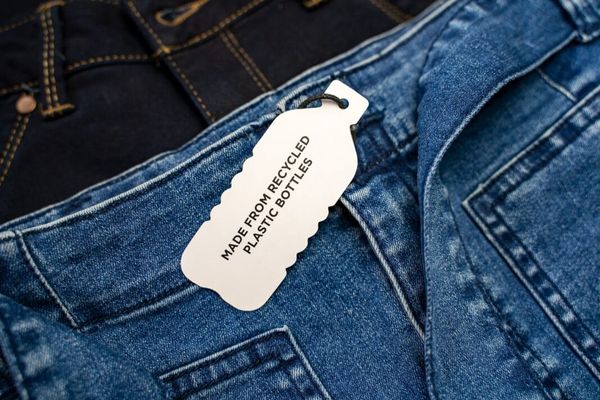 A close shot of a pair of denim jeans. Attached is a white paper label in the shape of a plastic bottle that reads "Made from recycled plastic bottles"
