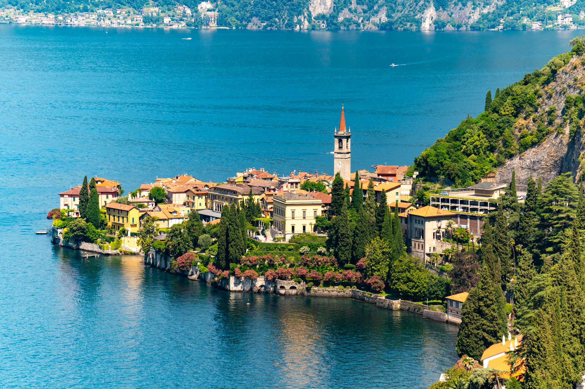 Summer aerial view of Varenna on Lake Como, showing the town's peninsula on the right and the panoramic landscape looking north up the lake.