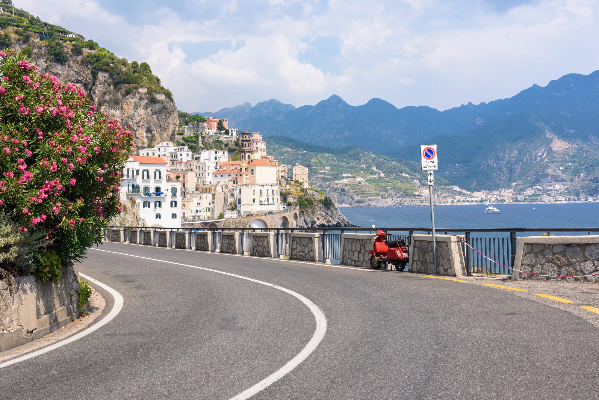 The famous, winding coastal road of the Amalfi Coast in Italy, with a classic red scooter parked alongside, offering breathtaking views of the turquoise Mediterranean Sea below.
