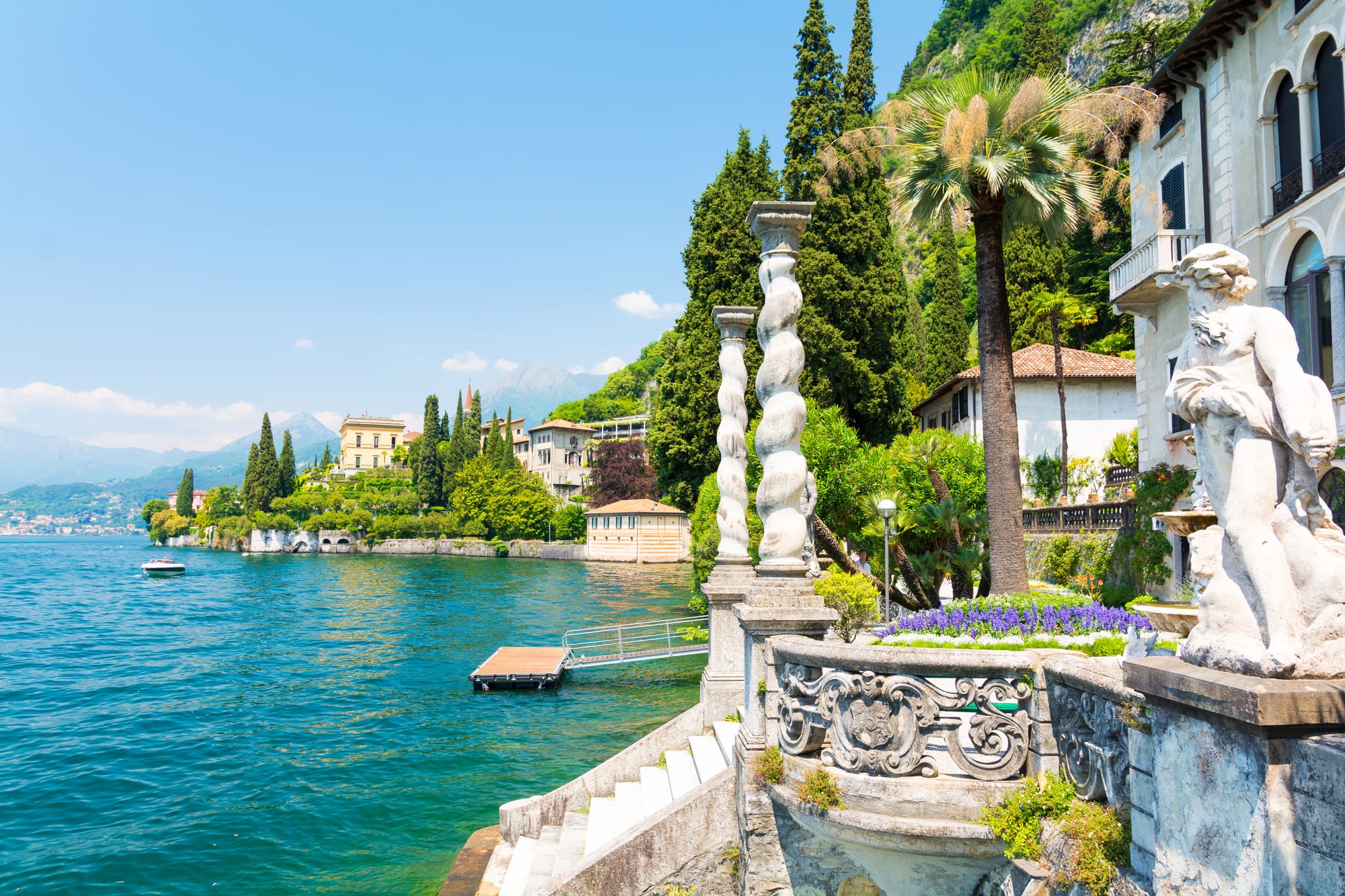 A glimpse of the Villa Monastero gardens in Varenna, featuring a classical statue and stone wall overlooking Lake Como on a sunny day.