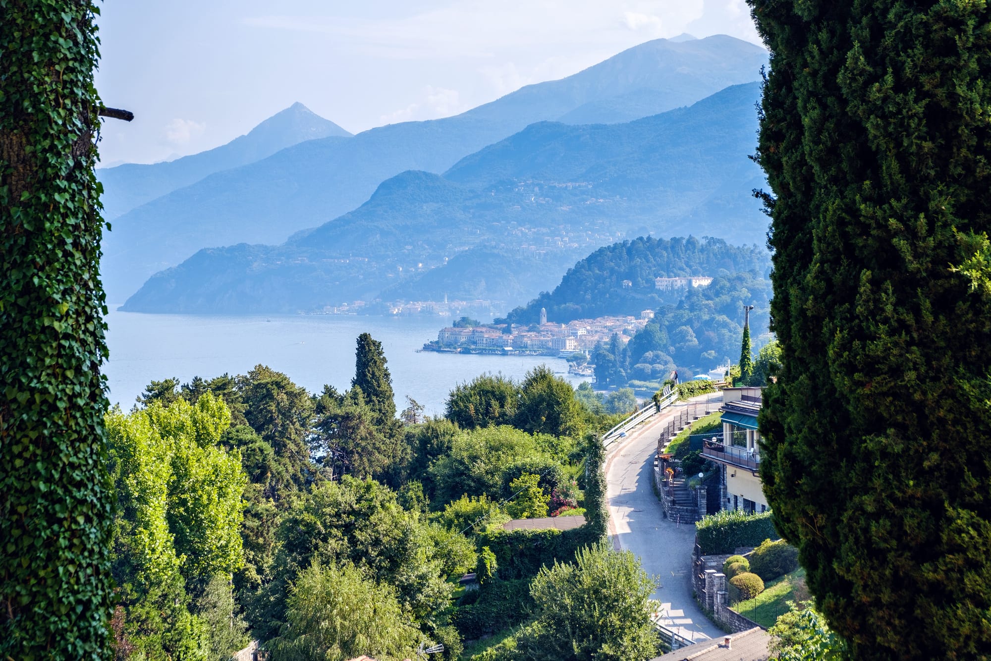 A winding coastal road snaking along the shore of the famous Lake Como, Italy. The narrow, curving asphalt is flanked by lush green mountains on one side and the deep blue water of the lake on the other.