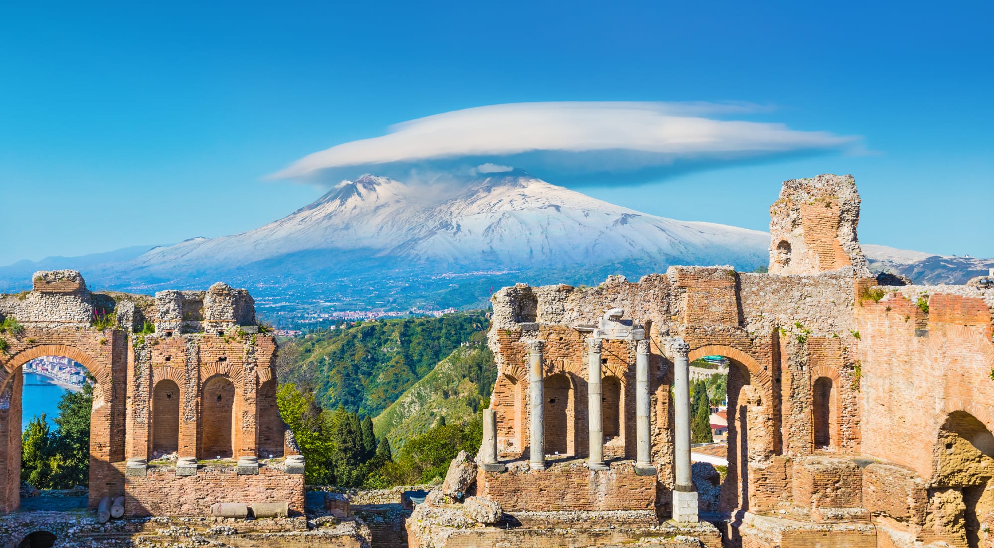 Panoramic view of the Greek Theater in Taormina, Sicily, with the ancient columns in the foreground and a smoking Mount Etna in the background.