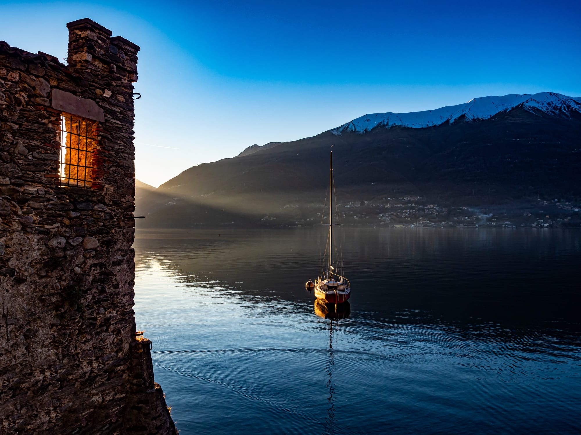 A single sailboat is moored in the tranquil waters of Lake Como at sunset, with the warm light illuminating the ancient medieval village of Corenno Plinio in the background.