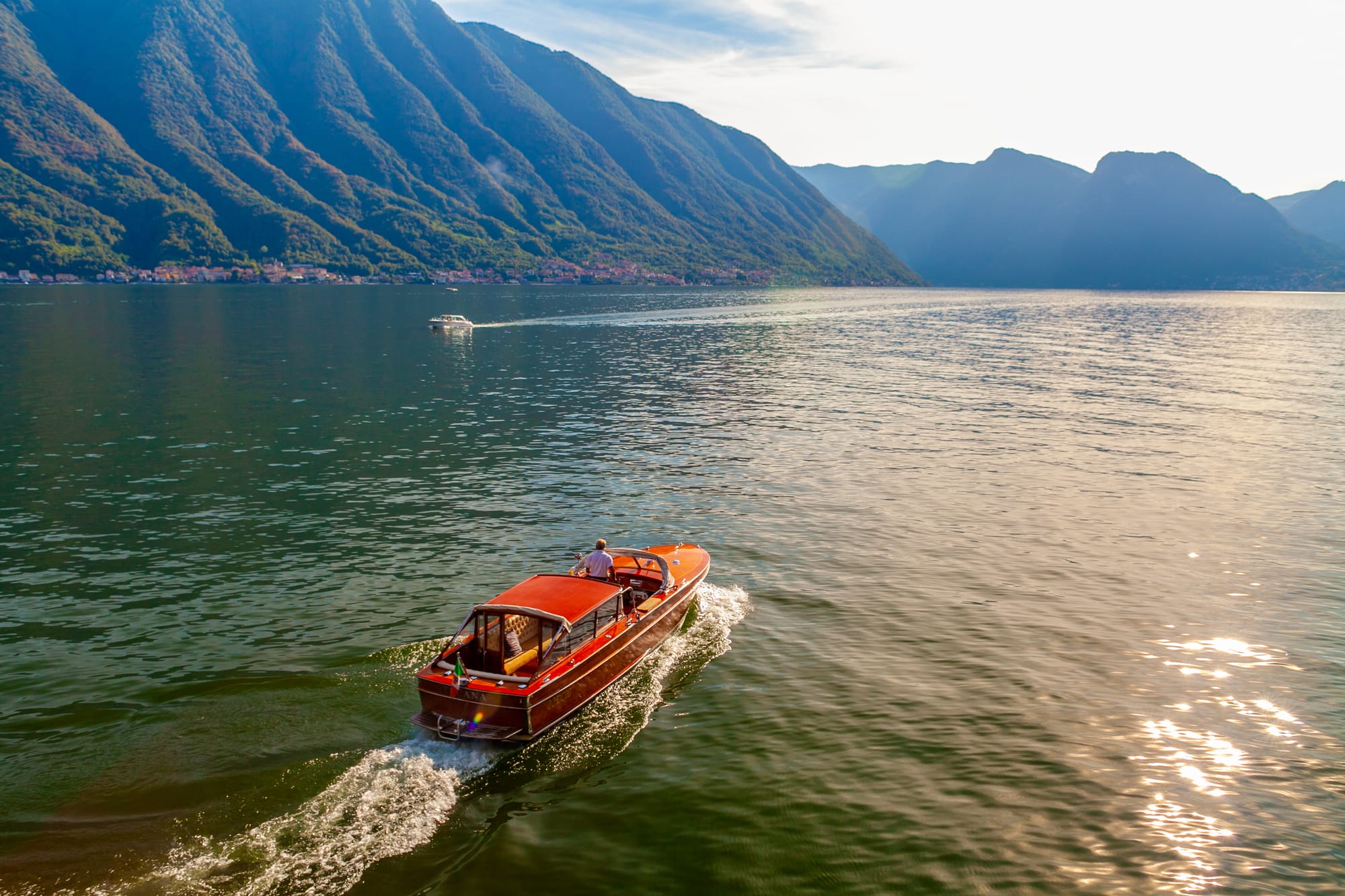 A classic luxury wooden Riva boat cruises on the calm blue water of Lake Como in September, with the shoreline of northern Italy in the background.