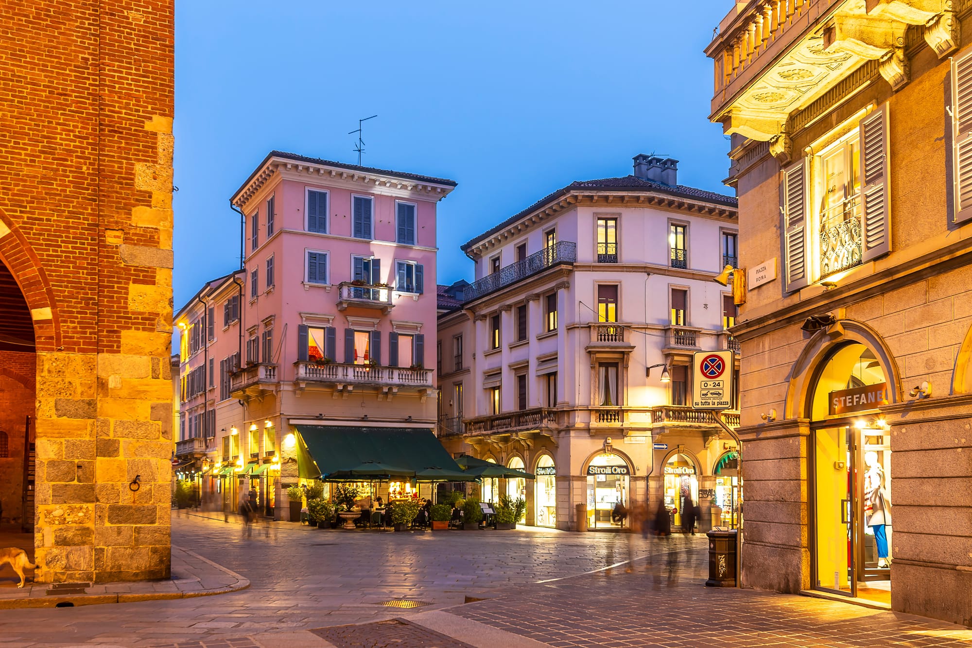 A charming cobblestone street in the historic center of Monza at dusk, with warm, inviting lights illuminating the historic buildings and storefronts.