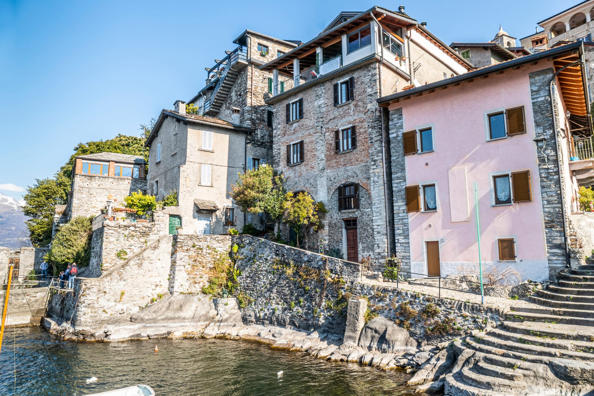 The waterfront of Corenno Plinio, where authentic stone houses with traditional Italian roofs meet the clear, blue water of Lake Como on a sunny day.