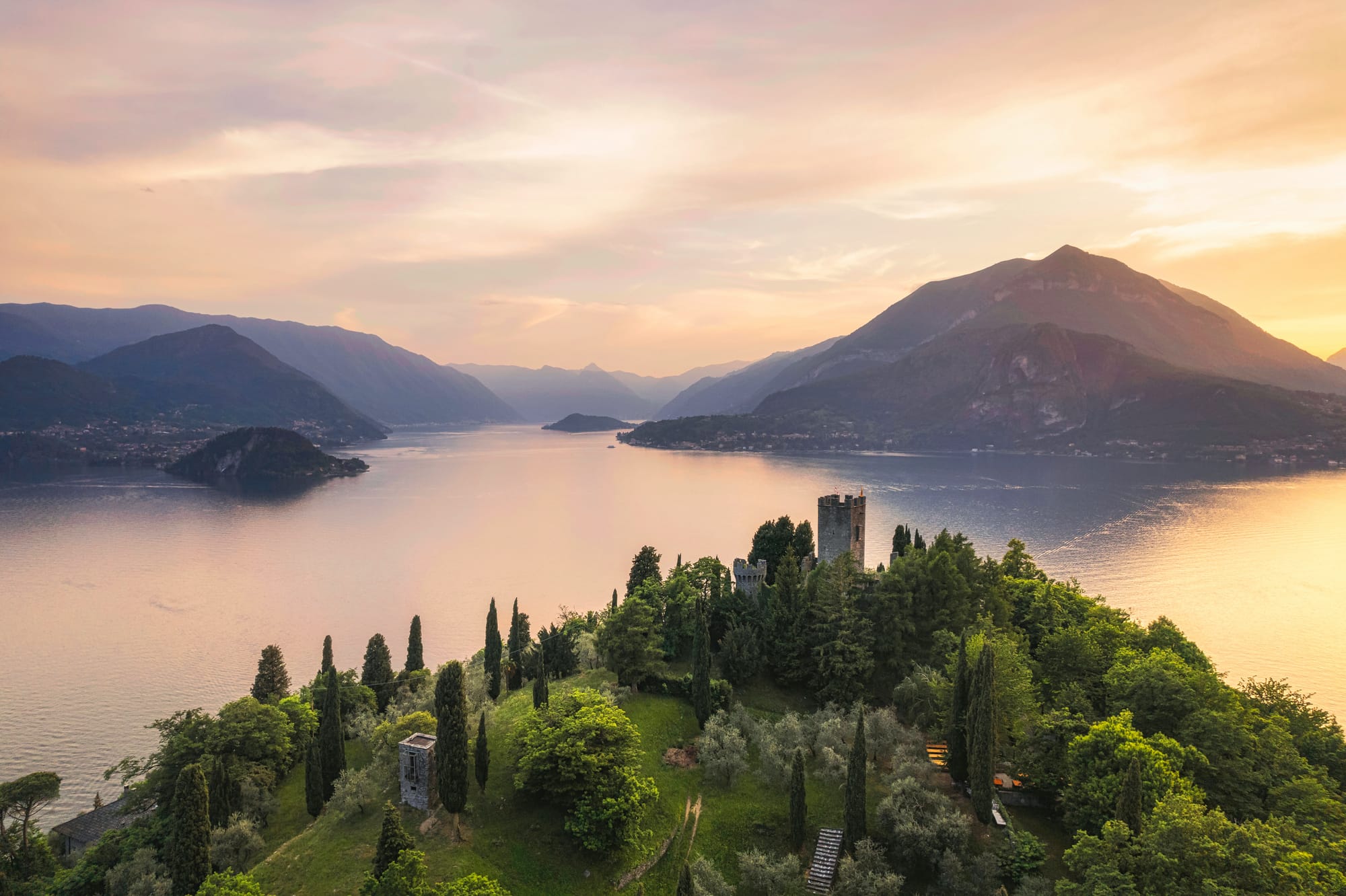 Stunning vertical aerial panorama of the medieval Vezio Castle high above the picturesque village of Varenna on Lake Como, Italy, captured by a drone at sunset with the Alps in the background.