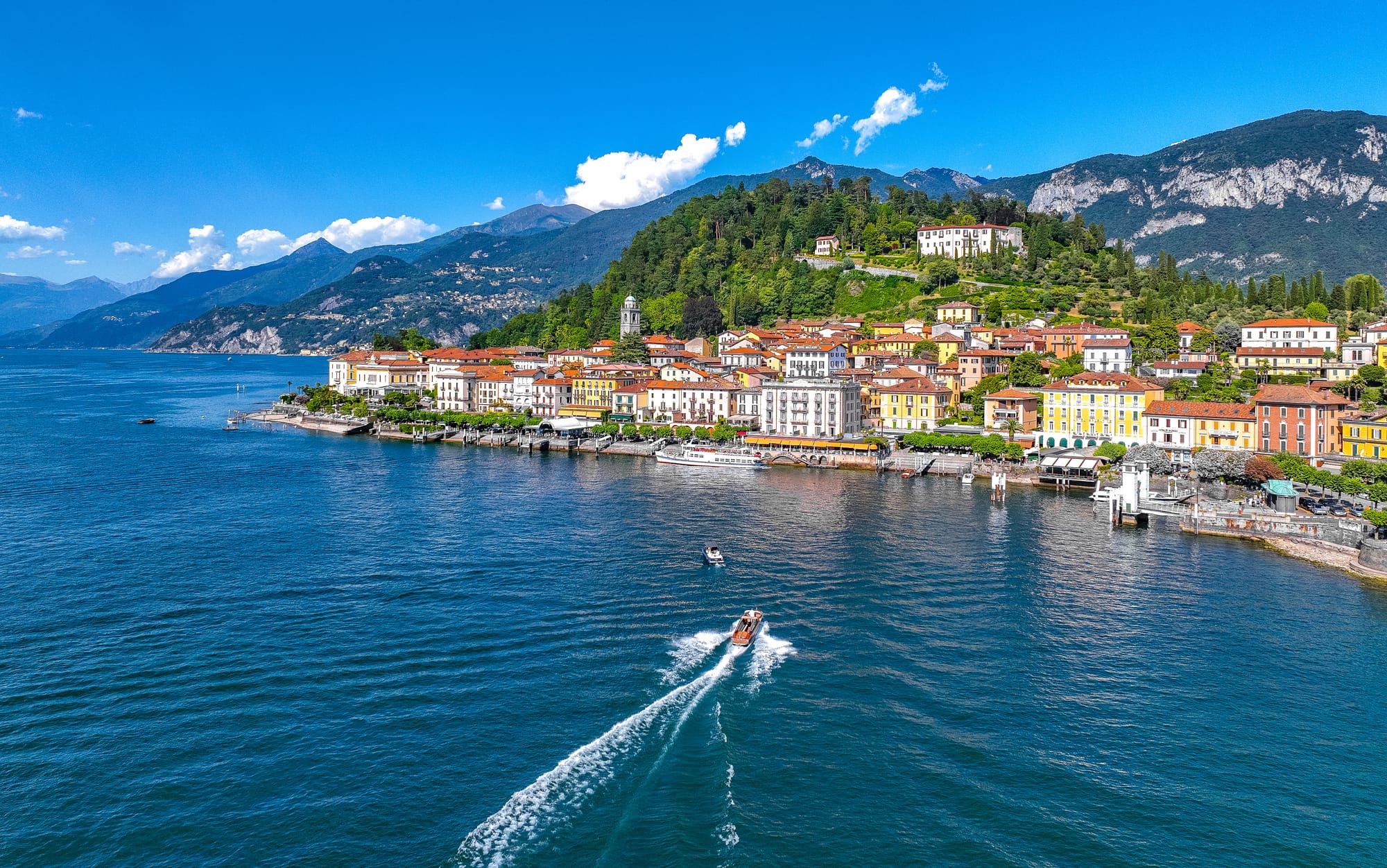 Aerial view from the water of the town of Bellagio on its promontory on Lake Como, with a boat approaching the shore.