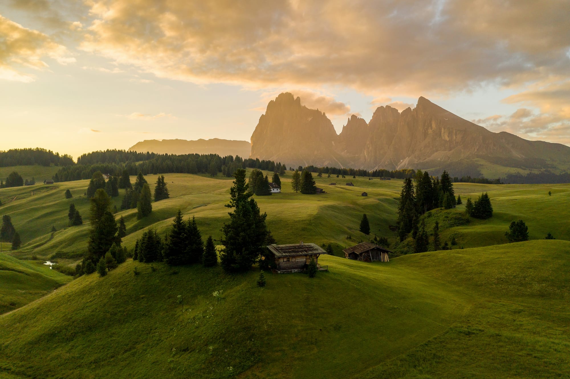 The rolling hills of Alpe di Siusi, the largest high-altitude Alpine meadow in Europe, bathed in the warm, golden hour light, with traditional wooden huts dotting the landscape.