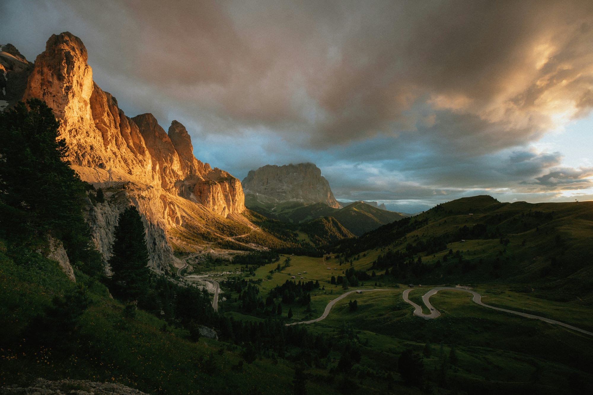 A scenic road winding through a valley in the Dolomites, Italy, at sunset, with the pale mountain peaks glowing pink in the evening light (Enrosadira). A highlight of the Great Dolomites Road.
