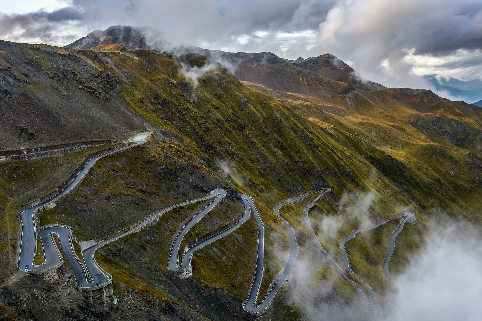 Incredible aerial view of the Stelvio Pass in the Italian Alps. The image shows the iconic series of hairpin turns as the road snakes up the steep, rocky mountain.