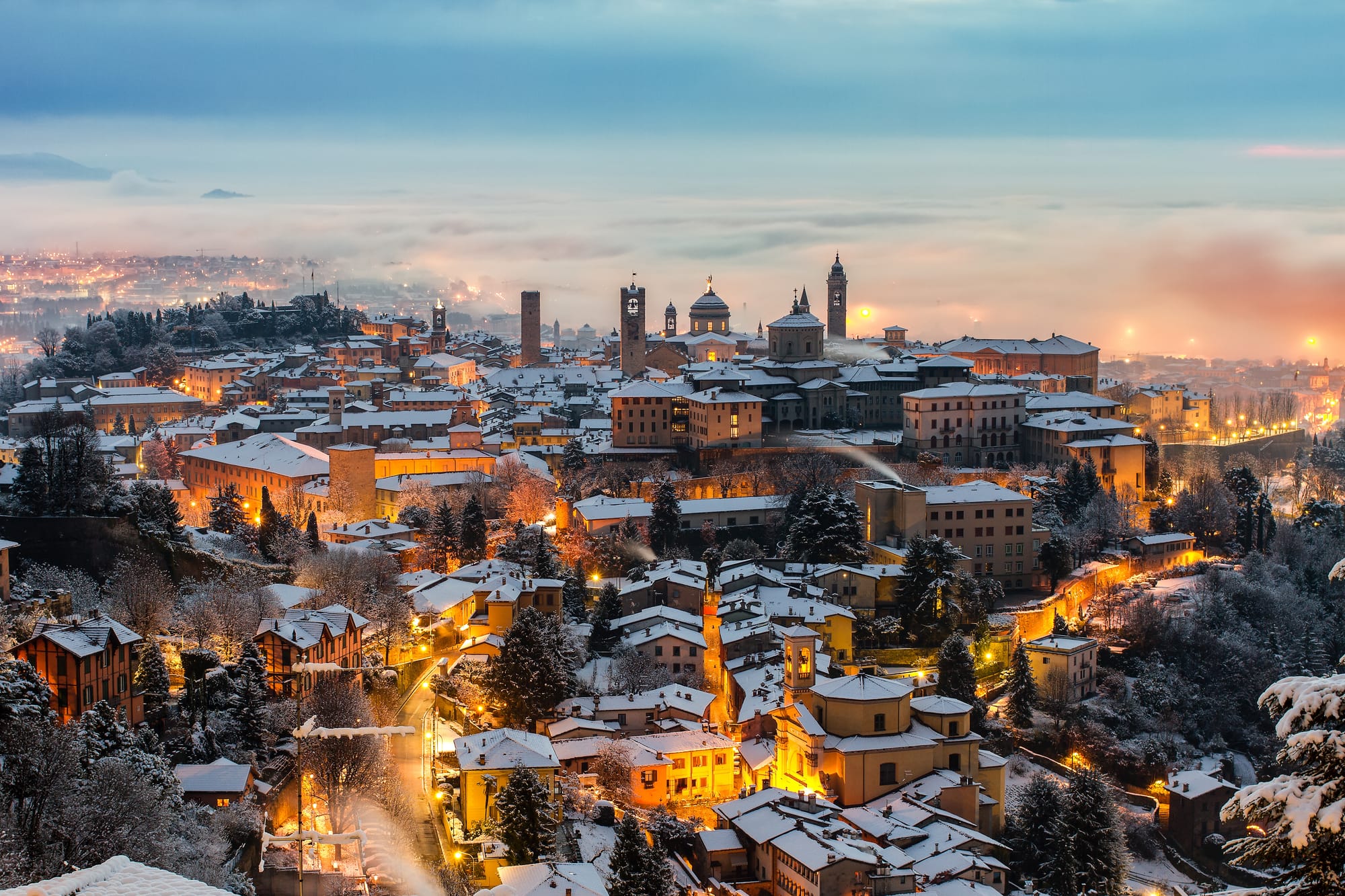 A panoramic winter evening view of Bergamo's illuminated Città Alta as seen from the higher vantage point of San Vigilio hill.