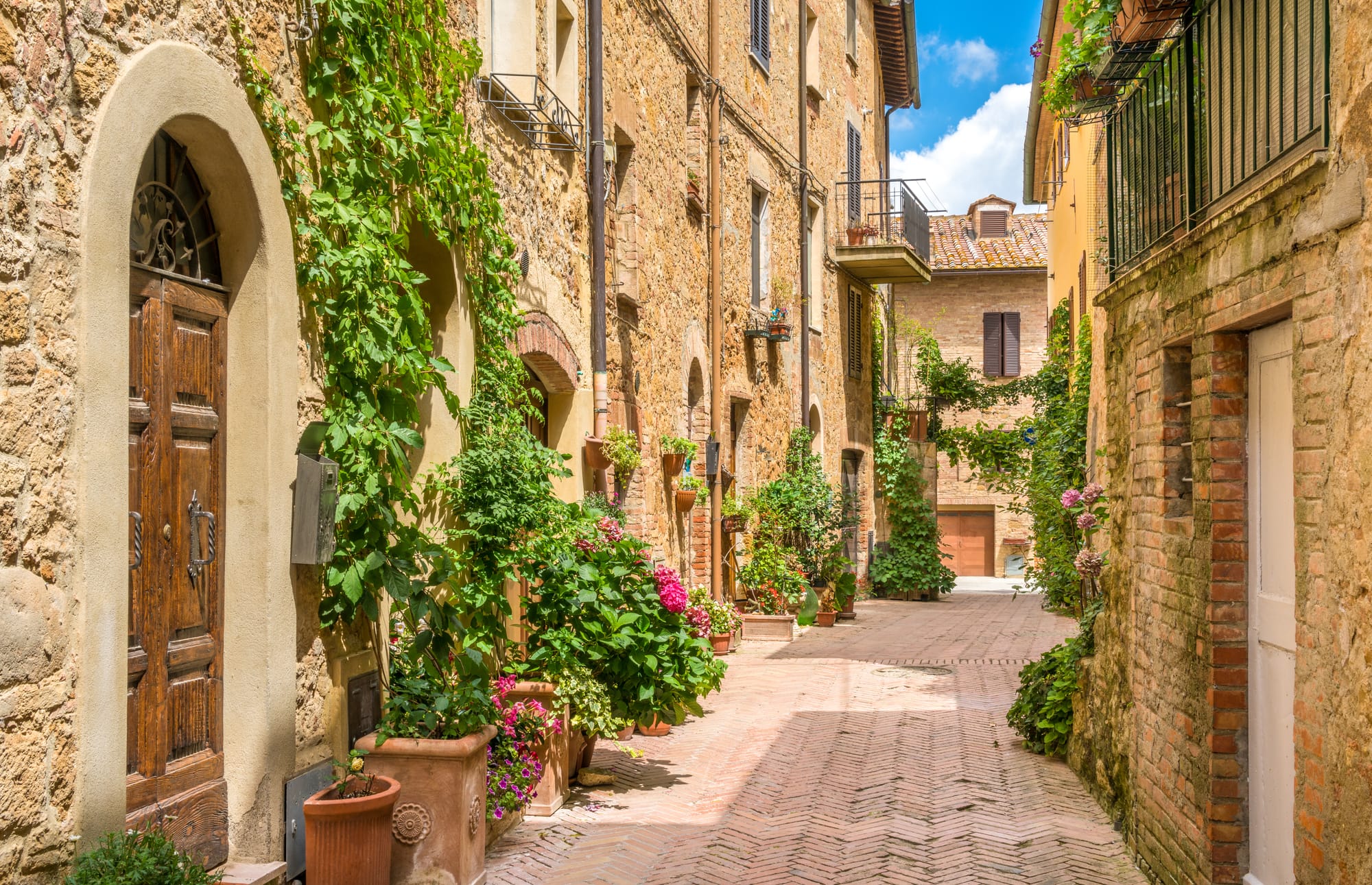 A narrow stone alley in Pienza, Tuscany, decorated with colorful flowers on the windowsills and doorways.
