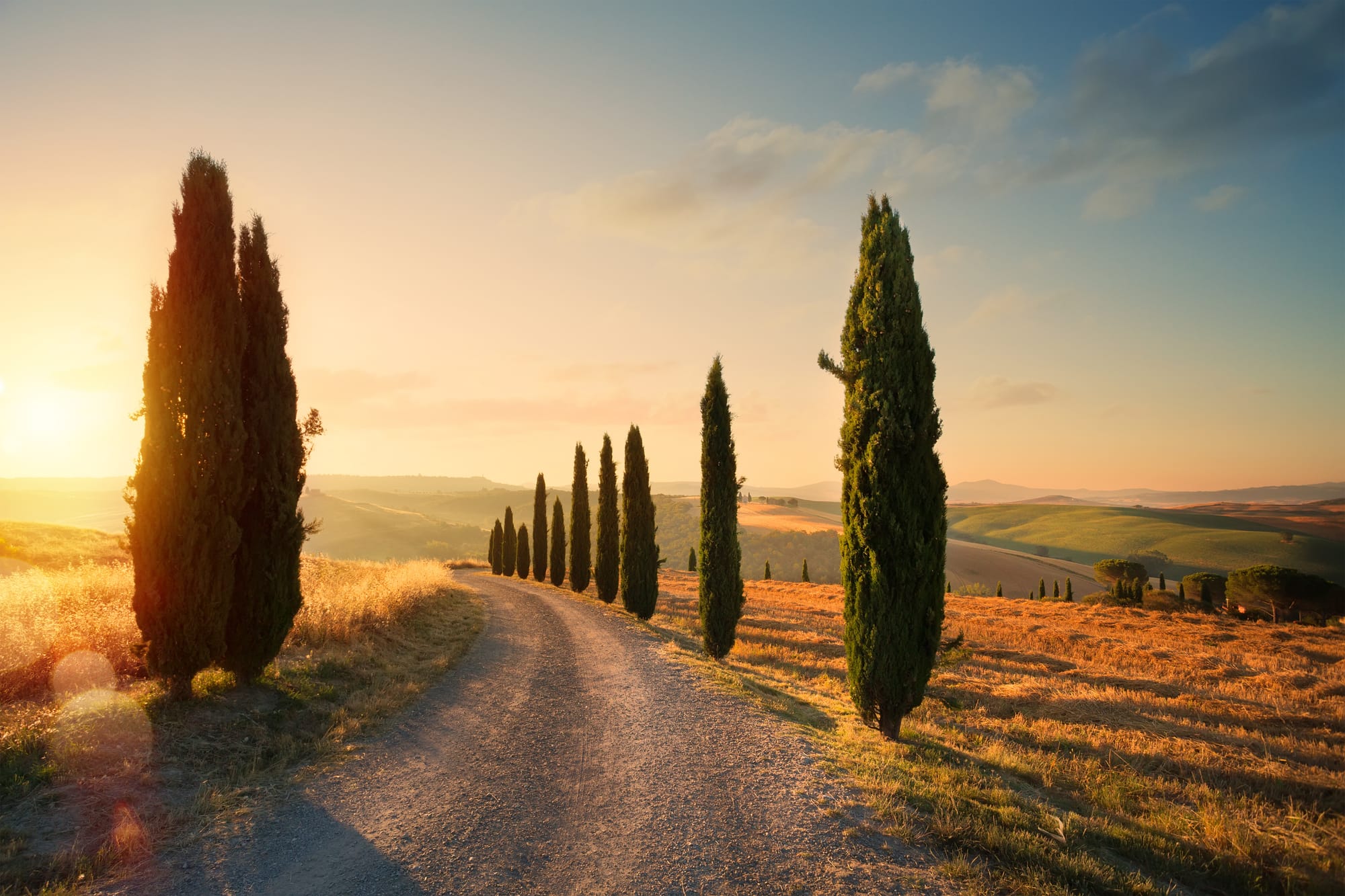 Golden sunrise light over the rolling hills of the Tuscan countryside in autumn, with a winding country road and misty valleys.