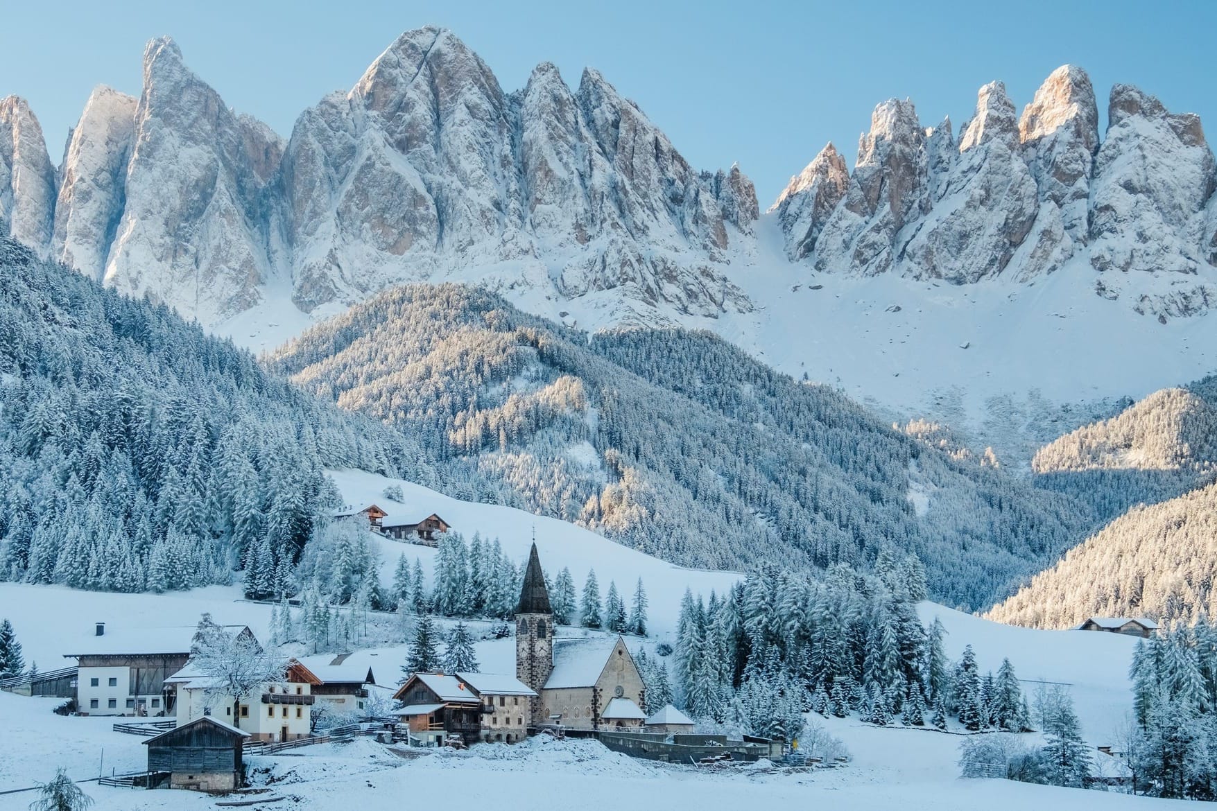 The small village in Val di Funes, Dolomites, covered in a thick blanket of snow during winter, with sharp mountain peaks in the background.