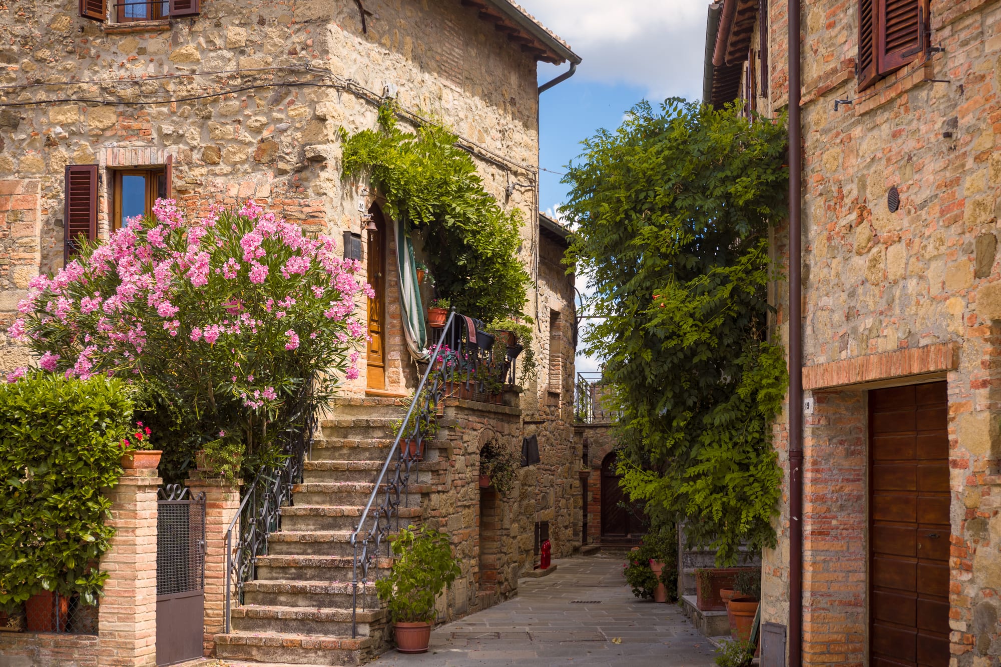 Beautiful medieval stone houses decorated with colorful flowers along a narrow cobblestone street in the village of Monticchiello, Italy.