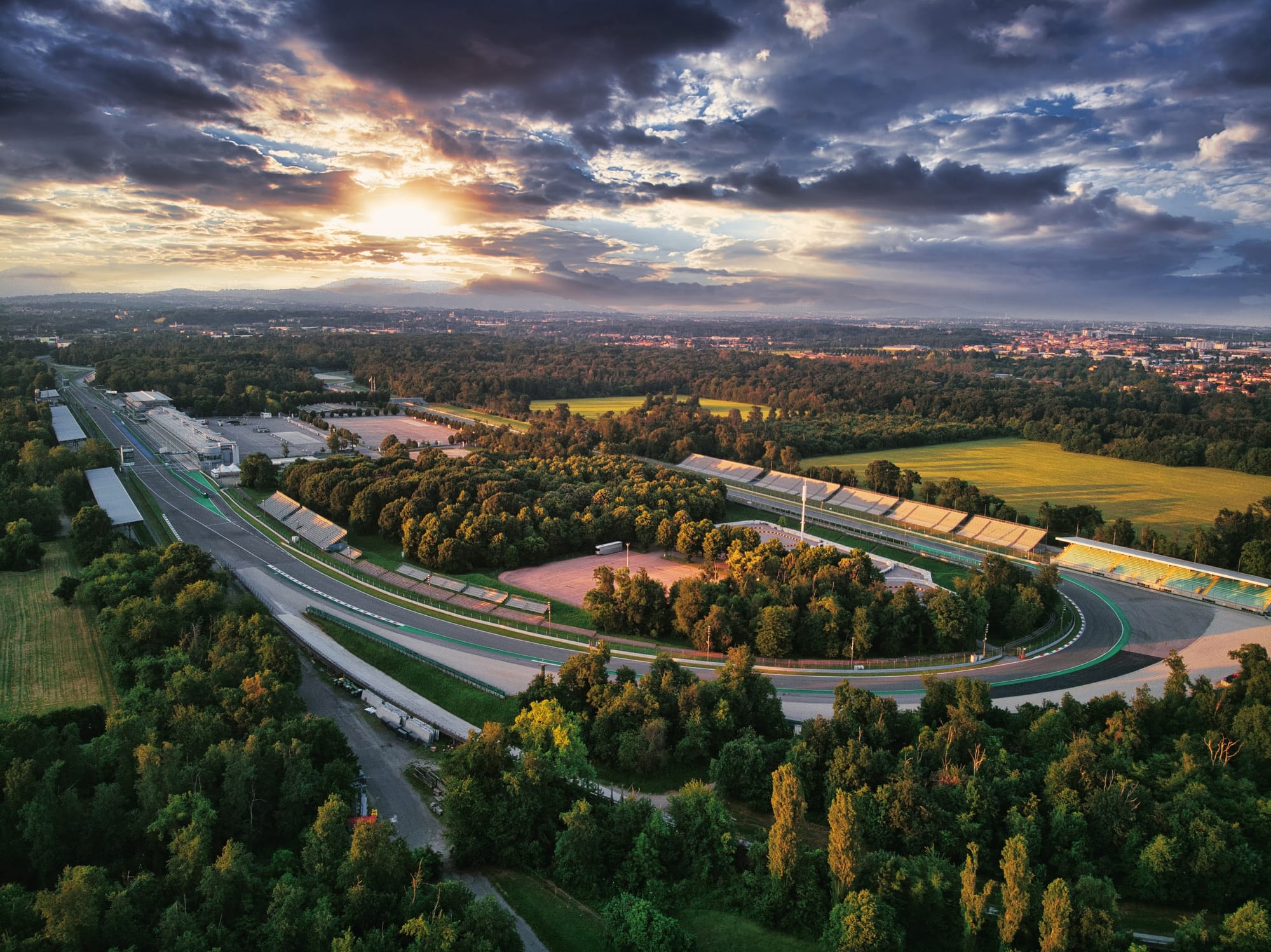 A stunning aerial photograph of the entire Monza circuit layout at sunset, nestled within the green trees of the Parco di Monza.