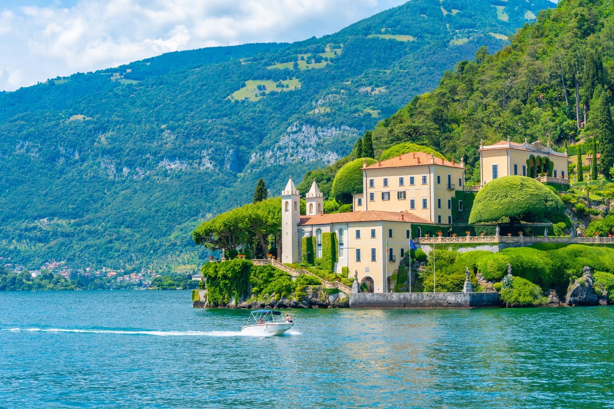 Exterior facade of a grand historic villa on the shores of Lake Como, showcasing neoclassical architecture and lush lakeside gardens viewed from a boat.