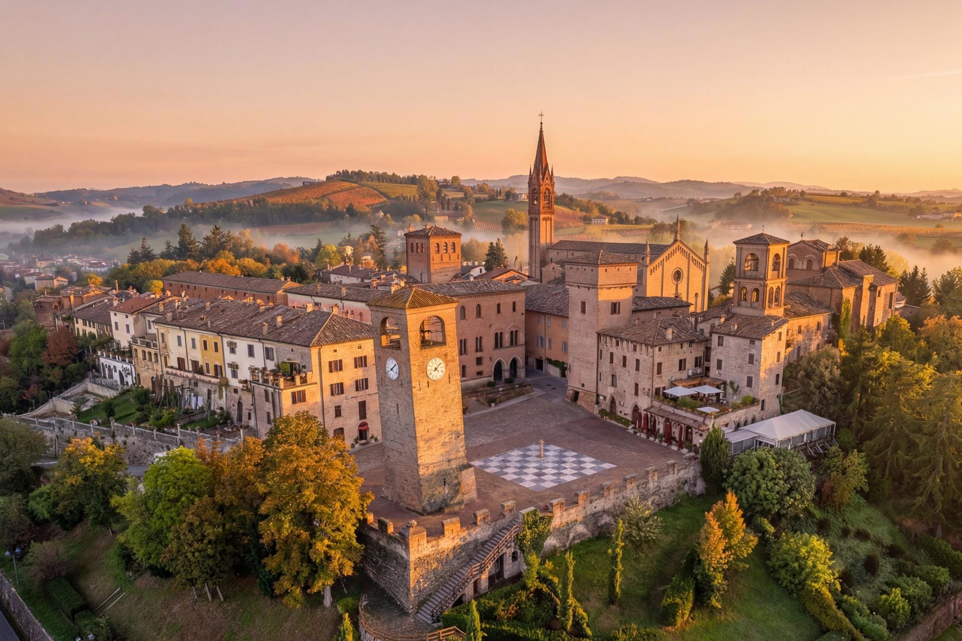 An aerial drone photograph of Castelvetro di Modena at sunrise, focusing on the checkerboard pattern of Piazza della Dama and the medieval towers surrounded by the rolling vineyards of the Food Valley.