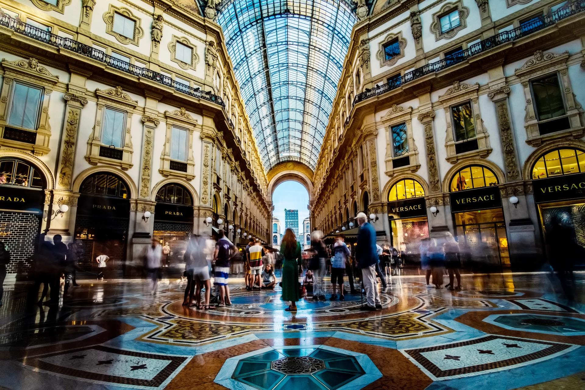 The ornate interior of the Galleria Vittorio Emanuele II in Milan, showing the historic glass dome and tourists exploring luxury boutiques.