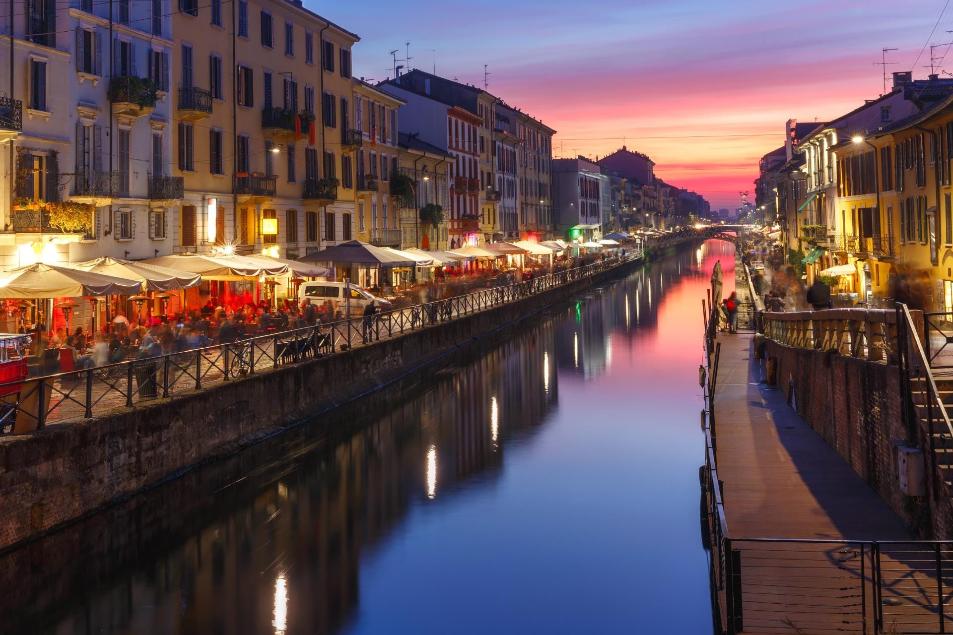 A scenic sunset view along the Naviglio Grande canal in Milan, with vibrant reflections in the water and cafes lining the pedestrian banks.