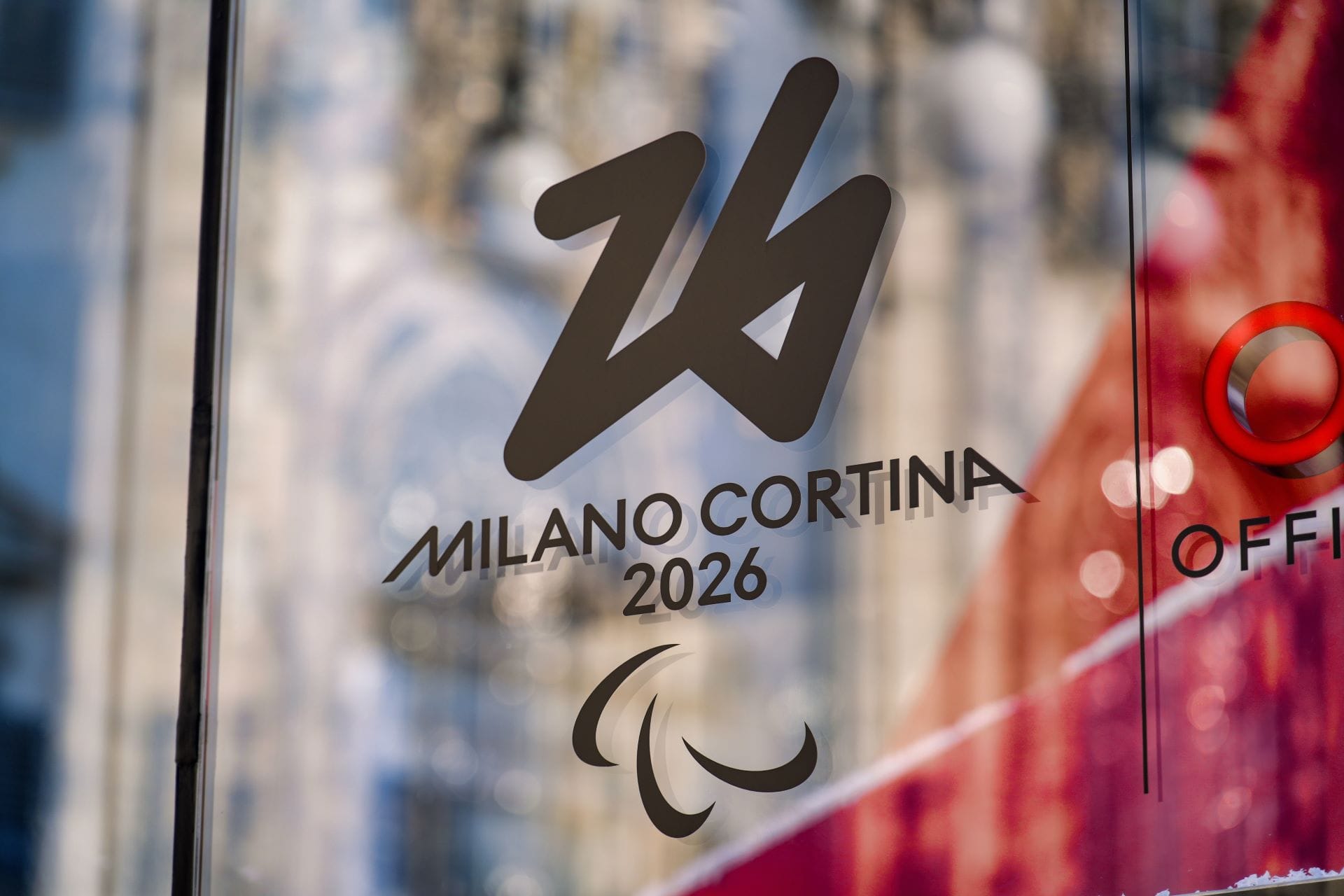 The Milano Cortina 2026 Olympic logo displayed on a glass window with a reflection of the Milan Cathedral (Duomo di Milano).
