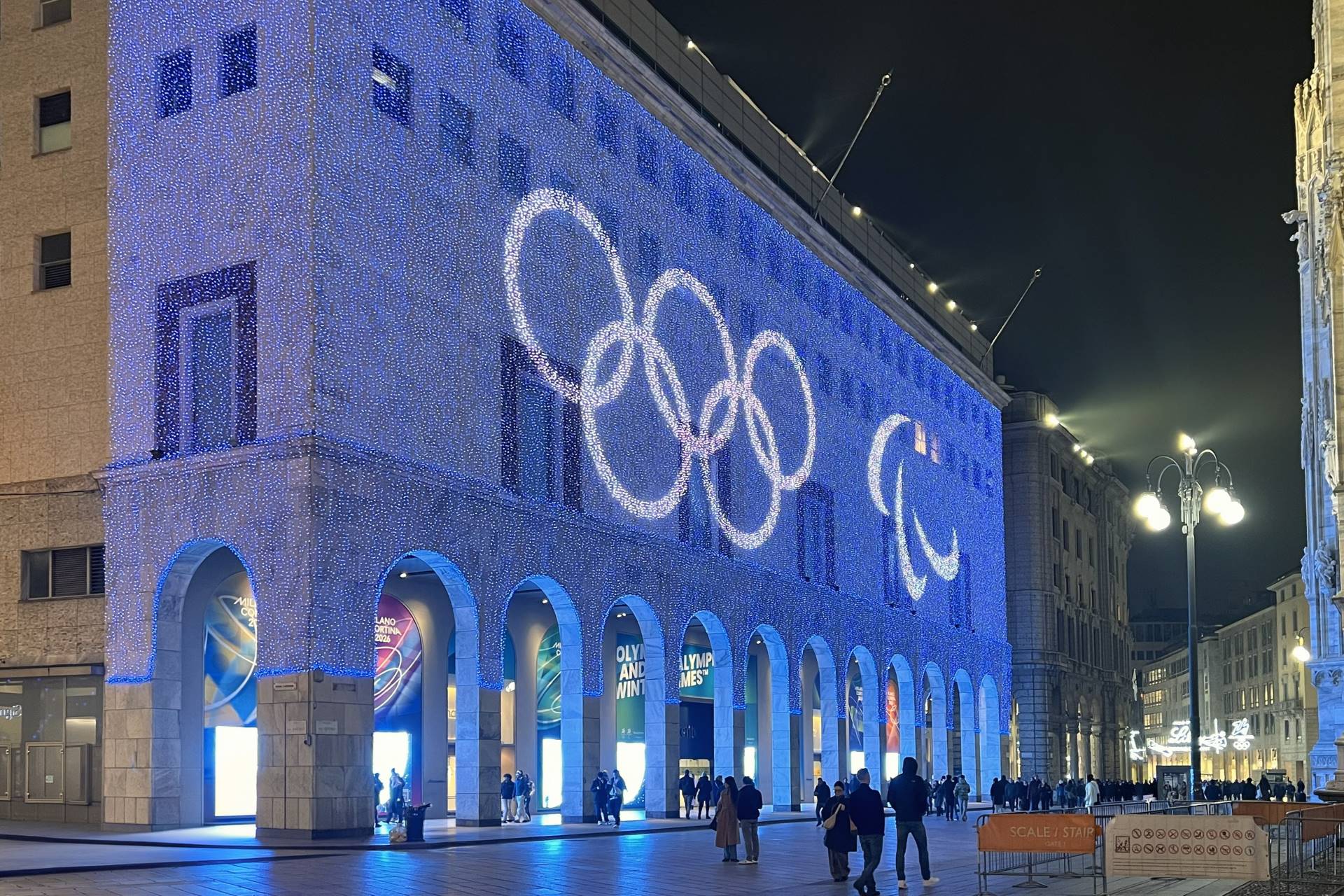 Illuminated olympic rings and paralympic agitos projected on the facade of a historic building in Piazza Duomo, Milan, with crowds gathering to celebrate the Milano Cortina 2026 winter games.