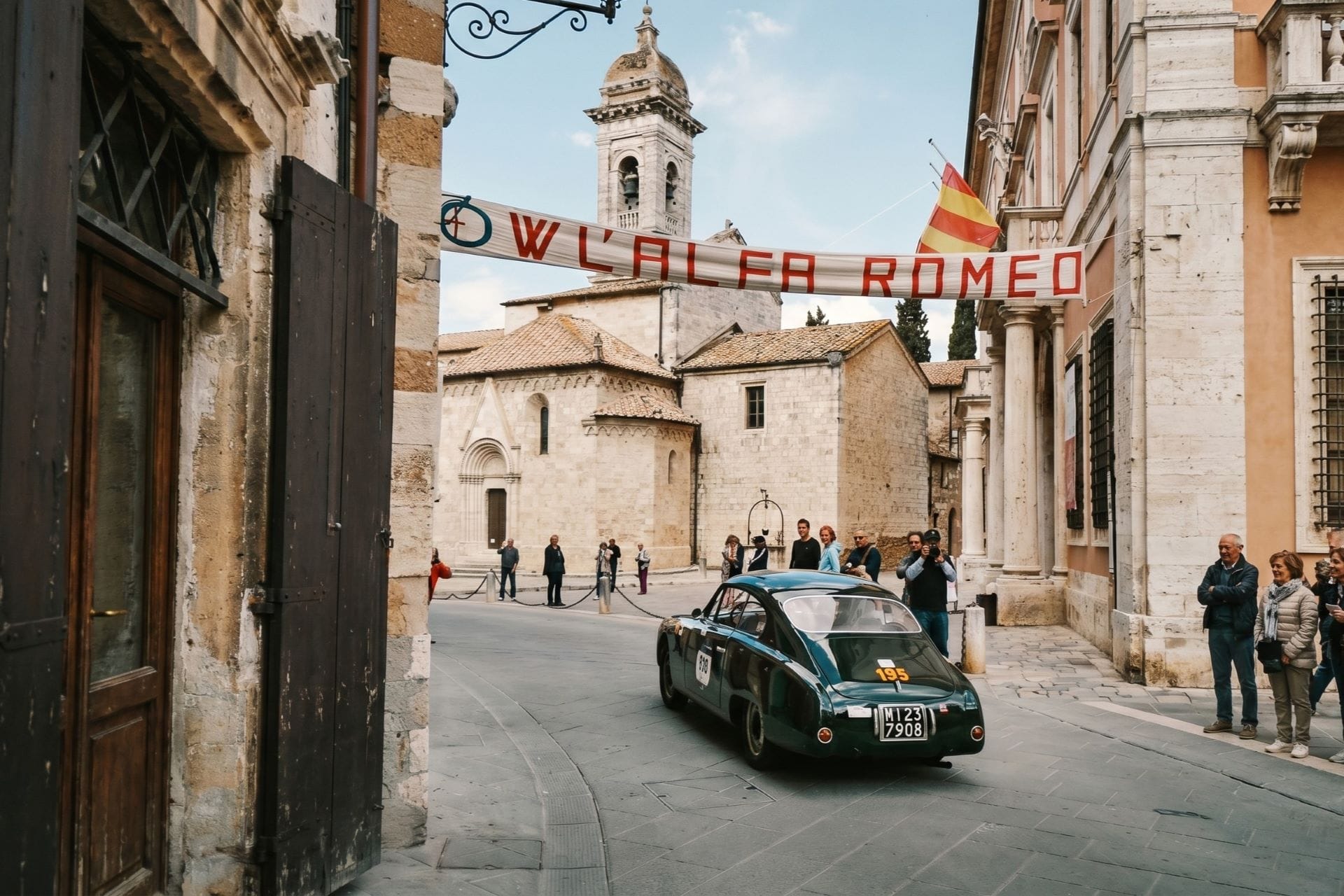 Dark green vintage coupe driving through a stone town square under an Alfa Romeo banner with spectators watching.