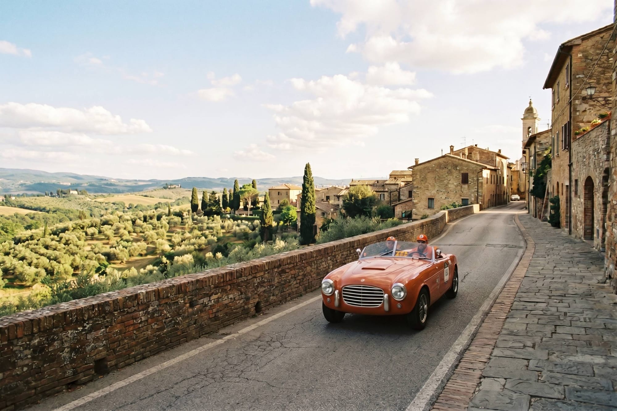 Drivers in a red vintage sports car waving to spectators along a stone village wall overlooking an olive valley.
